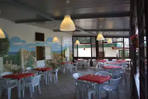 Überdachte Camping-Terrasse mit roten Tischen und weißen Stühlen, Wandgemälde und großen Fenstern im Hintergrund.