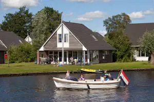 Haus mit Terrasse am Campingplatz am See, Ruderboot im Vordergrund, Rasen und Bäume im Hintergrund.