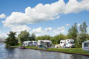 Campingplatz am Fluss: Wohnmobile und Zelte auf dem Stellplatz, Sitzgruppen und Fahrräder am Ufer, Bäume im Hintergrund.
