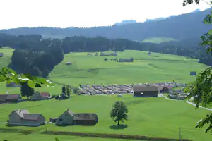 Campingplatz in grüner Berglandschaft mit Wohnwagen, kleinen Gebäuden und Wiesen, umgeben von Wäldern und Hügeln.