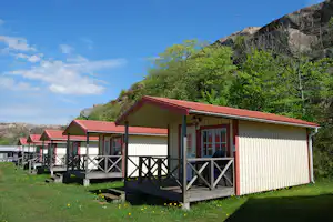 Ferienhaus mit rotem Dach und Holzterrasse, davor Stühle, im Hintergrund grüne Bäume und Felswand.