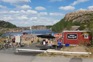 Campingplatz am Wasser mit roten Strandhütten, Sonnenschirmen, Fahrrädern und Blick auf Felsen, Bucht und bewaldete Ufer.