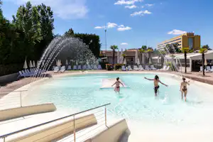 Schwimmbecken im Campingbereich mit Fontänen, Liegestühlen und Sonnenschirmen, im Hintergrund Bäume und ein Gebäude.