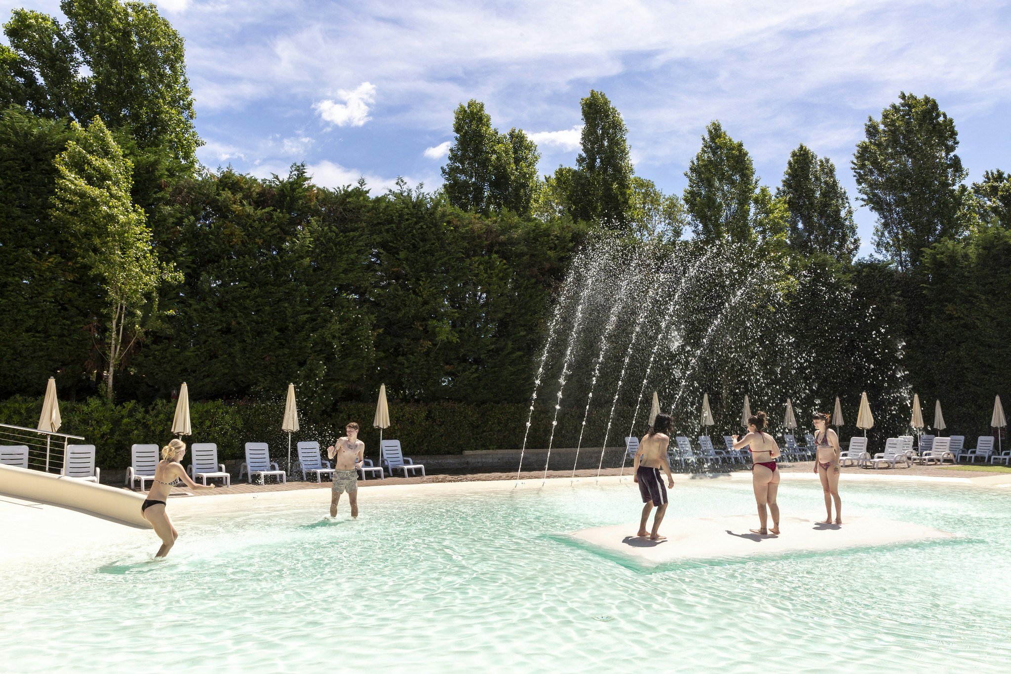 Schwimmbecken auf dem Campingplatz mit Wasserfontänen, Liegestühlen und Sonnenschirmen vor dichtem Grün und blauem Himmel.