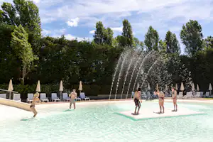 Schwimmbecken auf dem Campingplatz mit Wasserfontänen, Liegestühlen und Sonnenschirmen vor dichtem Grün und blauem Himmel.
