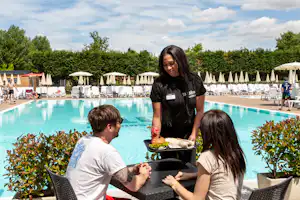 Poolbereich mit weißen Liegestühlen, Sonnenschirmen und einem Tisch am Becken, im Hintergrund Hecken und Gebäude.