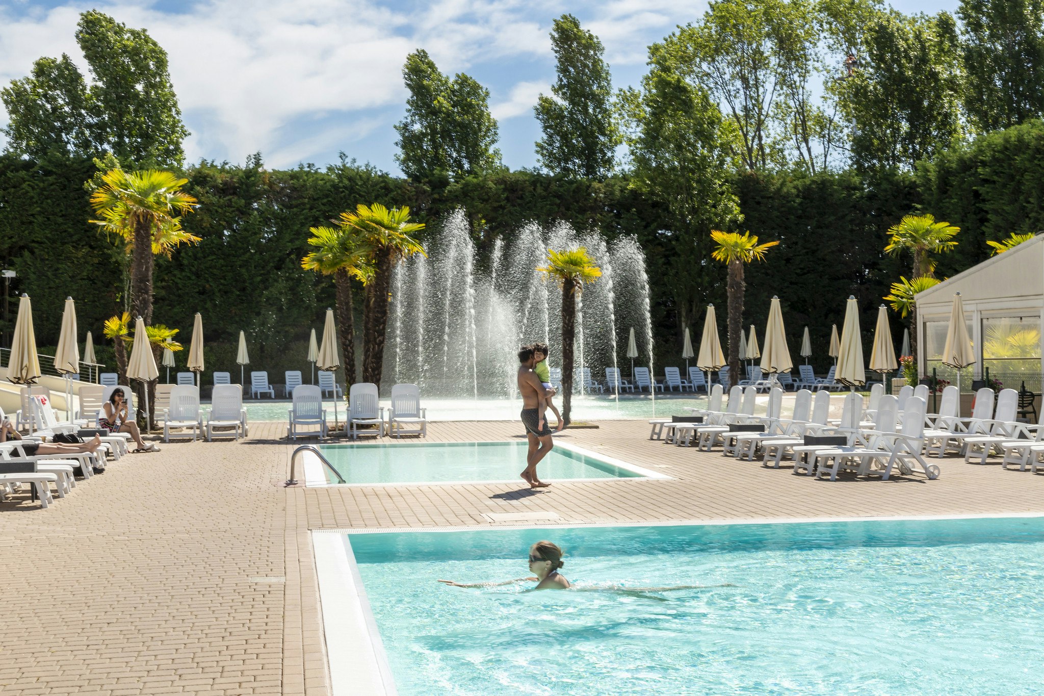 Schwimmbad mit Fontäne, Palmen und Liegestühlen, im Hintergrund hohe Bäume und Sonnenschirme am Campingplatz.