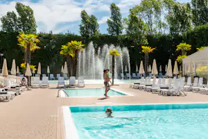 Schwimmbad mit Fontäne, Palmen und Liegestühlen, im Hintergrund hohe Bäume und Sonnenschirme am Campingplatz.