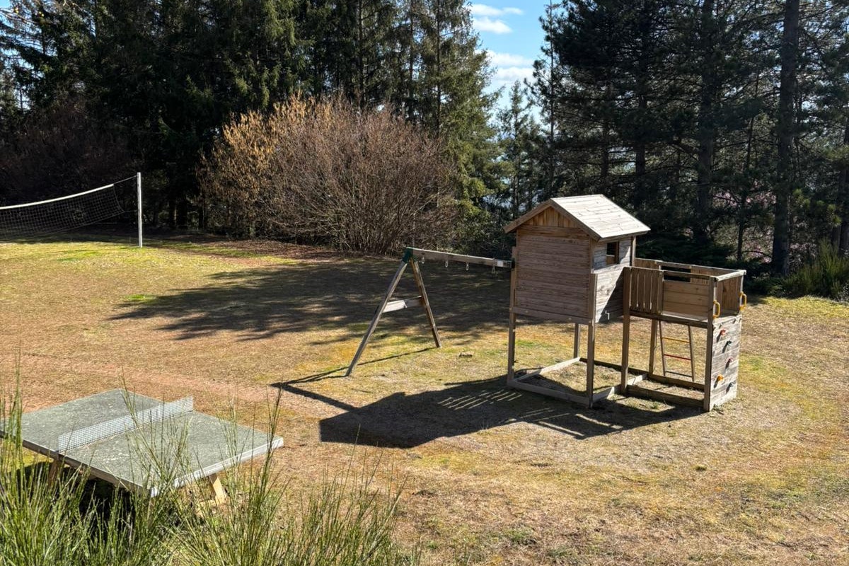 Holzhütte und Schaukel auf einem Campingplatz mit Rasen, Zaun und Baumreihe im Hintergrund.