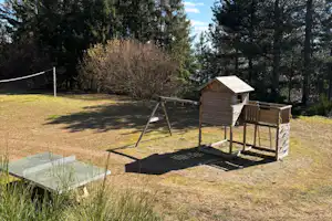 Holzhütte und Schaukel auf einem Campingplatz mit Rasen, Zaun und Baumreihe im Hintergrund.