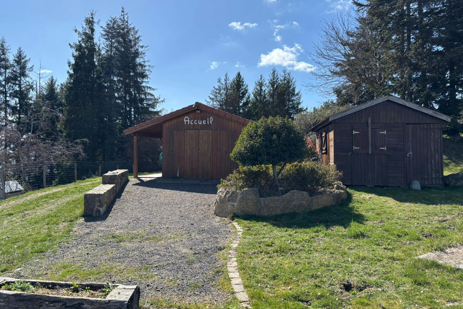 Holzunterstand und kleines Gebäude auf einem Campingplatz, mit Kiesweg, Rasenfläche und Bäumen im Hintergrund.
