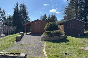 Holzunterstand und kleines Gebäude auf einem Campingplatz, mit Kiesweg, Rasenfläche und Bäumen im Hintergrund.