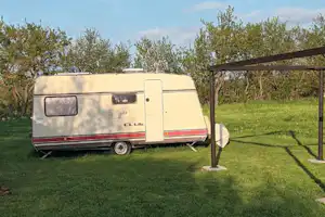Wohnwagen auf einem Campingplatz mit Vorzeltüberdachung, Rasenfläche und Büschen im Hintergrund.