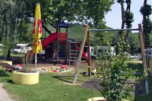 Spielplatz mit Rutsche und Klettergerüst im Campingbereich, daneben Wohnwagen und Bäume, Rasenfläche mit Sandspielzeug.