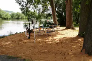 Terrasse am Fluss mit mehreren Stühlen und Tisch unter Bäumen, am Ufer mit Blick auf das Wasser.