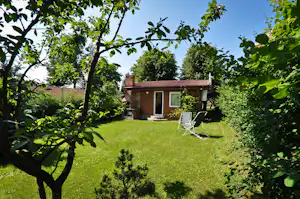 Kleines Gartenhaus mit Veranda und Schaukel im grünen Rasen, umgeben von Bäumen und Hecken.