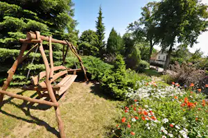 Holzrahmen im Campinggarten, umgeben von Blumenbeeten, Bäumen und einem Weg; im Hintergrund ein Gebäude.