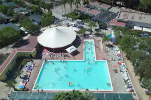 Luftaufnahme vom Campingplatz mit großem Pool, Sonnenschirmen, Liegen und Zeltpavillon, umgeben von Bäumen und Wohngebäuden.