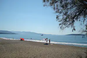 Sandstrand am See mit mehreren Liegestühlen, Sonnenschirmen und Boot in der Ferne, im Hintergrund Hügel und Wasserlinie.