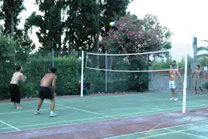 Tennisplatz auf dem Campinggelände mit Netz, Spielern und Hecke mit pinken Blumen, im Hintergrund Bäume.