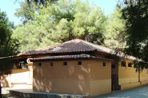 Campinggebäude mit Ziegeldach und mehreren Fenstern, umgeben von Bäumen und Grün im Hintergrund.