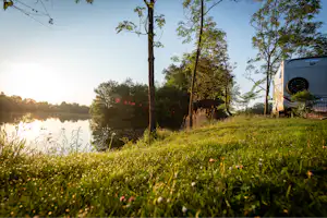 Campingzelt am Seeufer auf einer grünen Wiese, im Hintergrund Bäume und ruhiges Wasser bei Sonnenlicht.