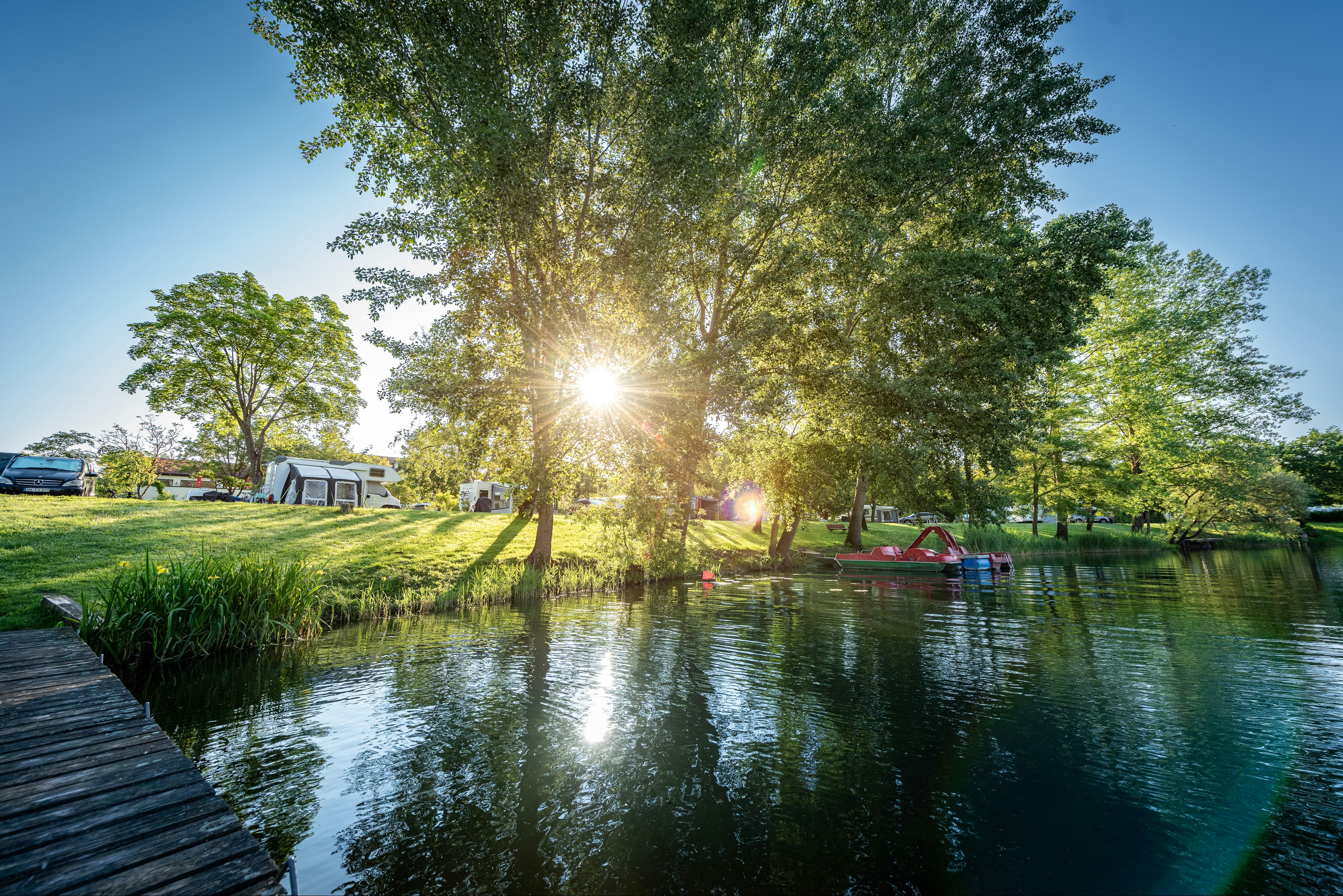 Ruhiger See am Campingplatz mit Bäumen, Sonnenlicht und einem Steg am Ufer.