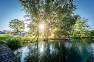 Ruhiger See am Campingplatz mit Bäumen, Sonnenlicht und einem Steg am Ufer.