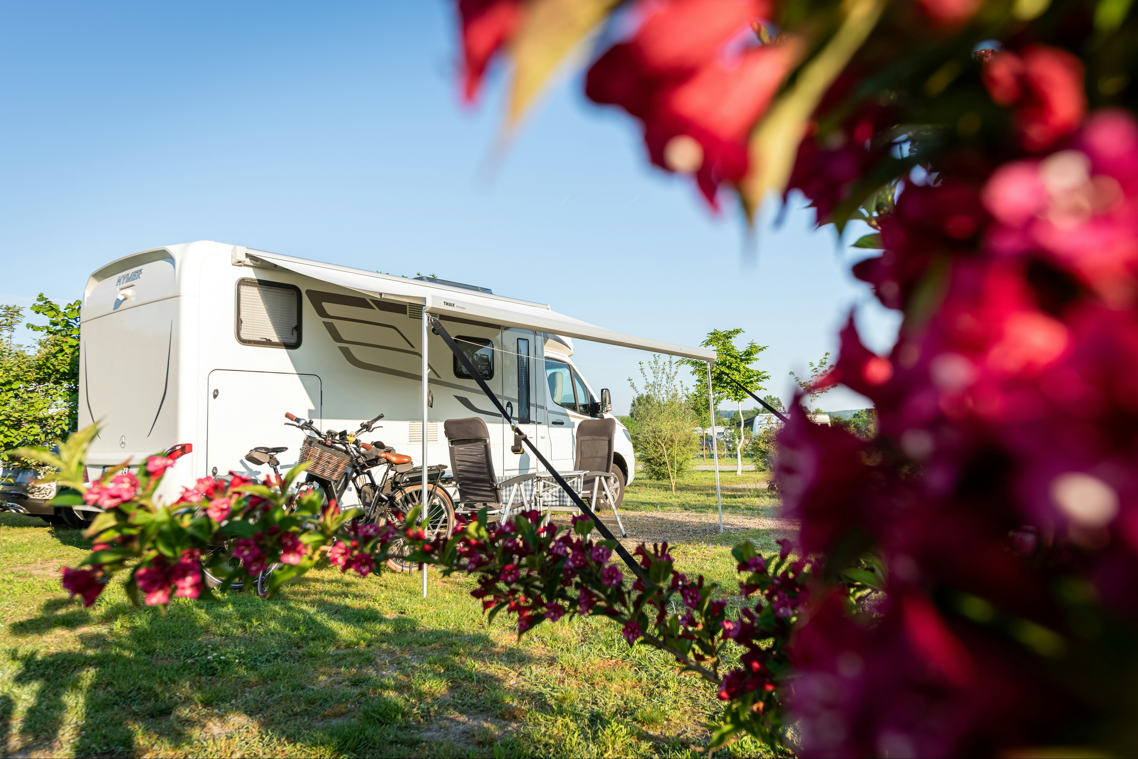 Wohnmobil auf einem Campingplatz, im Vordergrund Blumenbeete und Rasenfläche, im Hintergrund Bäume und ein Stellplatzweg.
