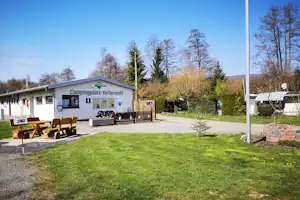 Gebäude mit Schild „Campingplatz Kellerwald“, Infotafel und Sitzbank vor gepflegtem Rasen, Bäume und Weg im Hintergrund.