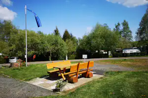 Holzbank auf Kiesweg am Campingplatz, daneben Fahnenmast mit blauem Banner, im Hintergrund Wohnwagen und Bäume.