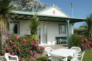 Bungalow mit überdachter Terrasse, weißen Gartenstühlen und Tisch, umgeben von blühenden Sträuchern und Palmen im Campingbereich.