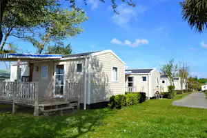 Reihen von Mobilheimen auf einem Campingplatz, mit Holztreppe, Veranda und Rasen, umgeben von Bäumen und blauem Himmel.