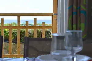 Blick von der Camping-Terrasse mit Holzgelaender, Tisch und Weinglaesern; im Hintergrund ist das Meer zu sehen.
