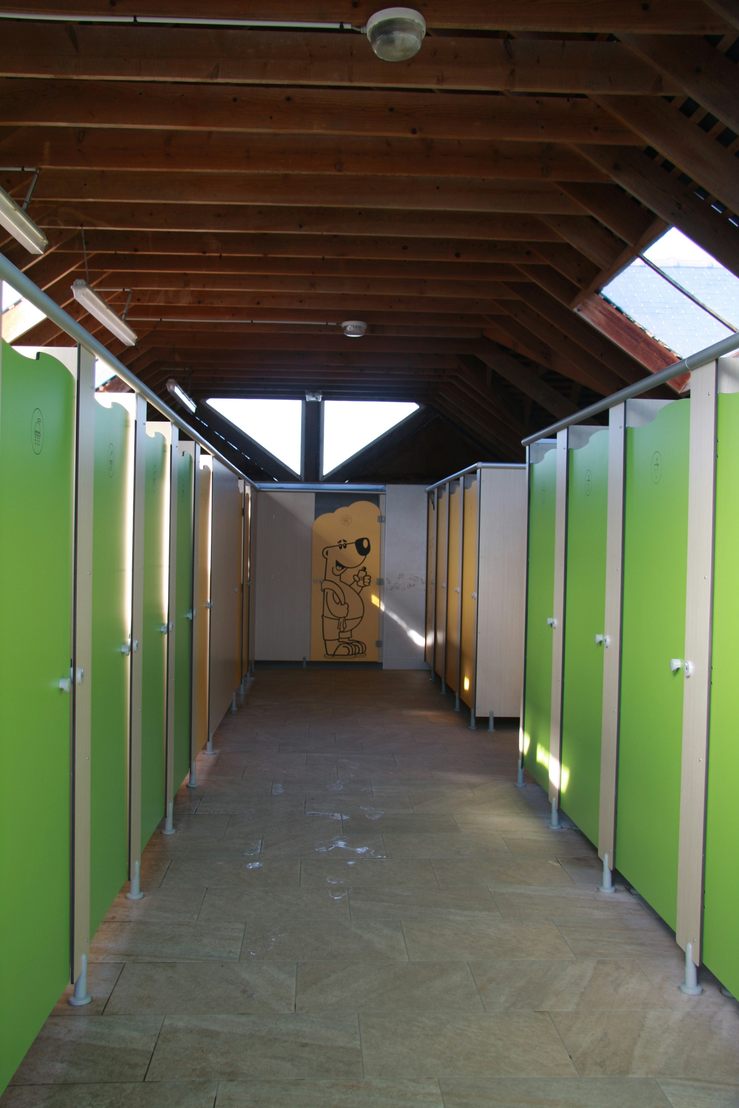 Gang unter Holzdecke mit grünen Türen, grauem Boden und beleuchtetem Durchgang, im Hintergrund eine Wand mit Zeichnung.