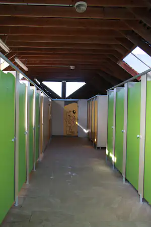 Gang unter Holzdecke mit grünen Türen, grauem Boden und beleuchtetem Durchgang, im Hintergrund eine Wand mit Zeichnung.