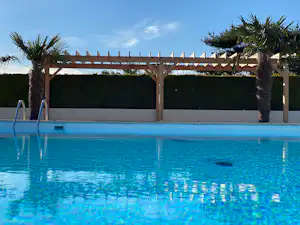 Blauer Camping-Pool mit Leiter, Palmen und Holzpergola am Rand, dahinter Hecke und klarer Himmel.
