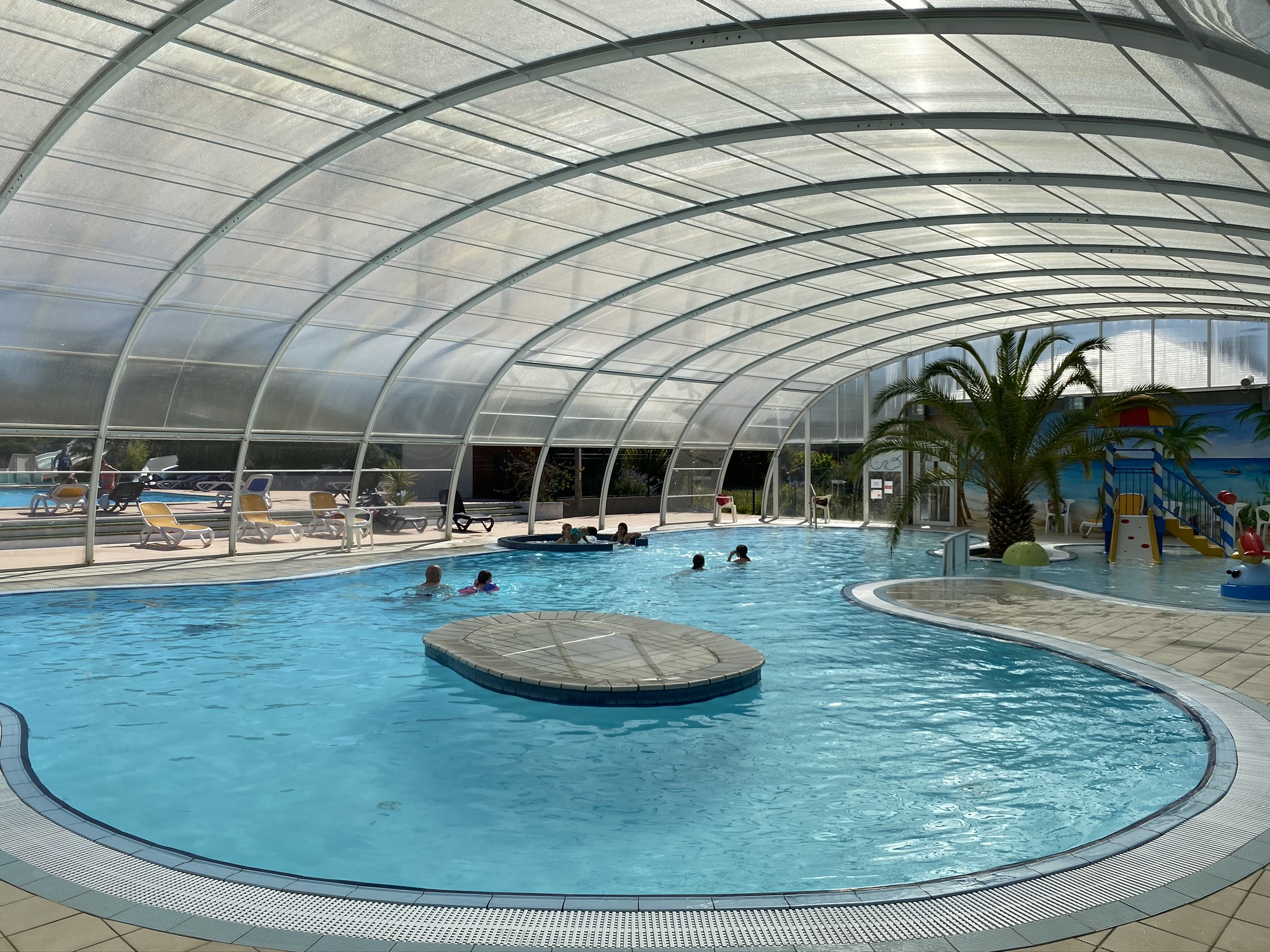 Überdachtes Schwimmbecken mit rundem Sprungbereich, Pooldach aus Glas und Liegen am Beckenrand.