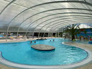 Überdachtes Schwimmbecken mit rundem Sprungbereich, Pooldach aus Glas und Liegen am Beckenrand.
