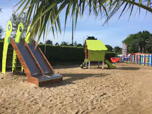 Sandiger Spielplatz mit brauner Rutsche, bunten Spieltischen und Palmenzweigen im Hintergrund.