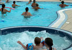 Familie im Schwimmbecken eines Campingplatzes, Kinder und Erwachsene planschen im Wasser, im Hintergrund ein weiteres Becken.
