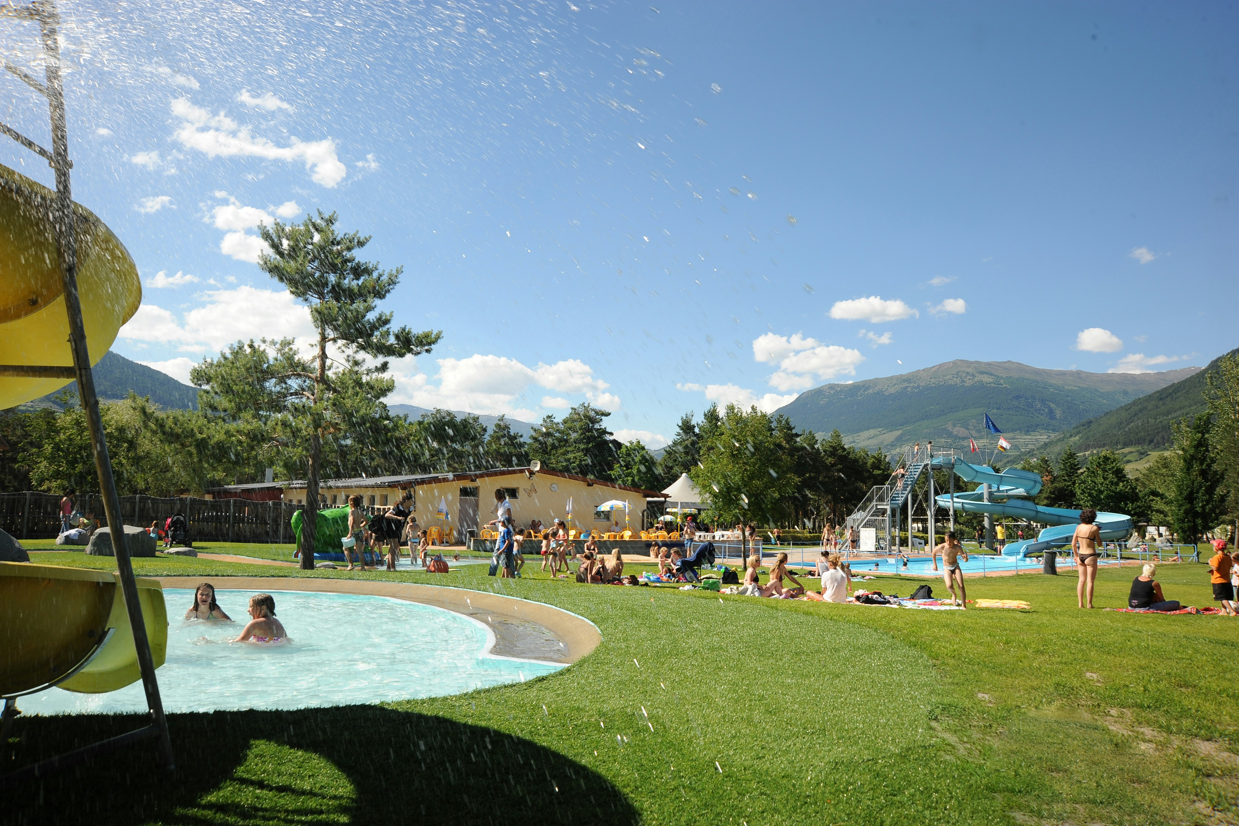 Schwimmbecken mit Rutsche und Liegen am Campingplatz, grüner Rasen, Bäume und Berge im Hintergrund.