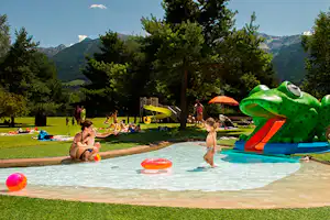 Campingpool mit Wasserrutsche, Kinder planschen im Becken, bunte Schwimmreifen auf dem Wasser, Bäume und Berge im Hintergrund.