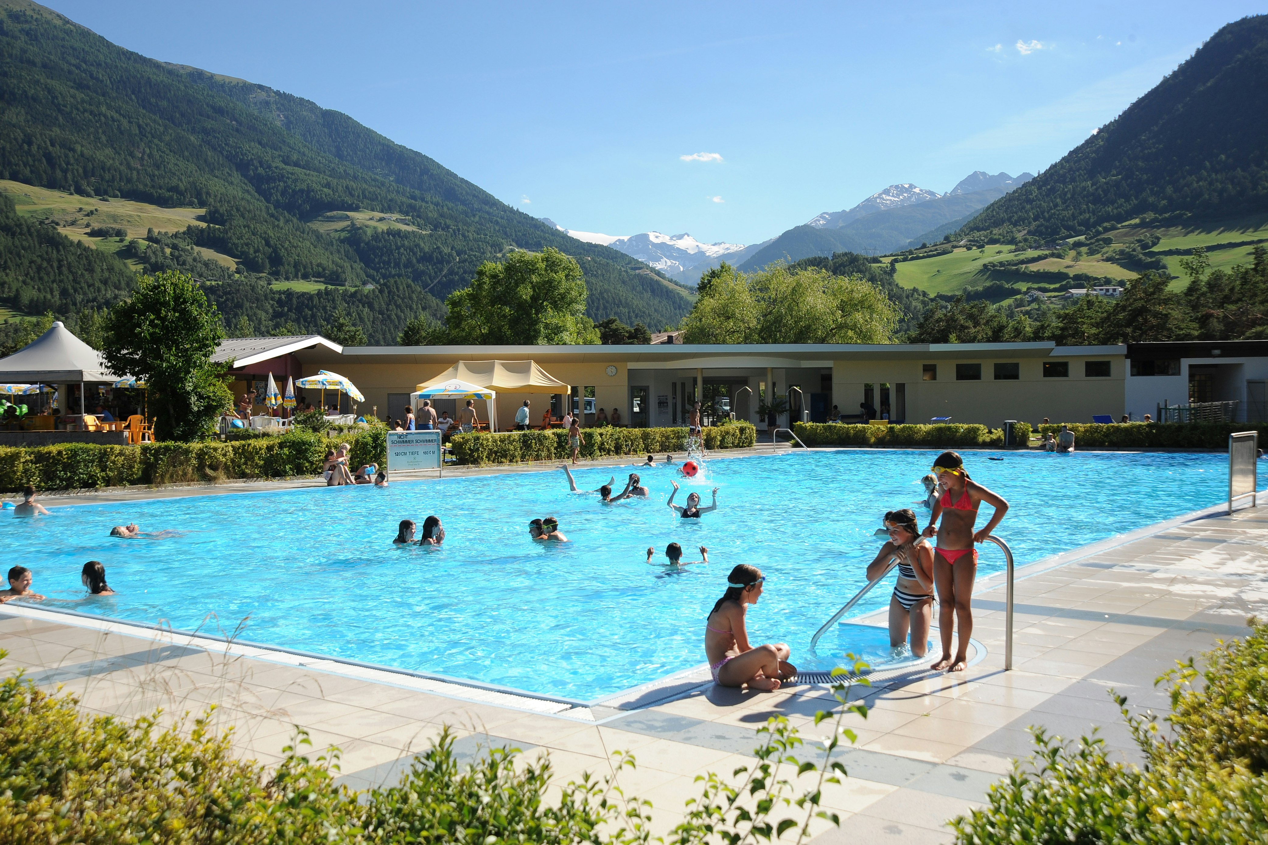 Großes Schwimmbecken im Campingbereich mit Menschen im Wasser, Liegen und Blick auf grüne Berge und Tal.