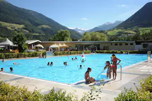 Großes Schwimmbecken im Campingbereich mit Menschen im Wasser, Liegen und Blick auf grüne Berge und Tal.