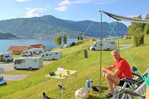 Camping Kjørnes- Camper sitzen vor dem Wohnmobil im Schatten mit Blick auf die Berge und auf den See