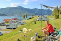 Camping Kjørnes- Camper sitzen vor dem Wohnmobil im Schatten mit Blick auf die Berge und auf den See