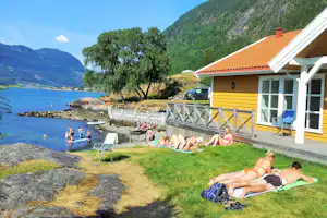 Gelbes Ferienhaus am See mit Terrasse, Grilltisch und Liegestühlen am Ufer, im Hintergrund Berge und Bäume.