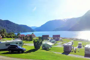Campingplatz am See mit Wohnhäusern, Stellplätzen und Wohnmobilen, umgeben von grünen Bergen und blauem Wasser.