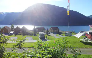 Campingplatz am See mit Zelten, kleinen Häuschen und Rasenfläche, im Hintergrund Berge und Wasser.